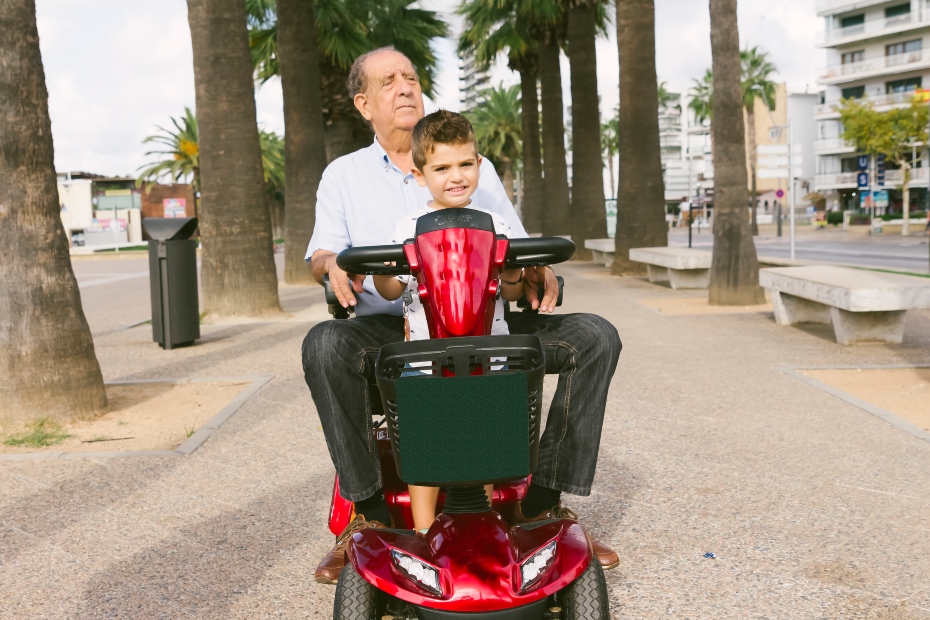 A grandfather with his grandson on his lap riding through town on his ruby red mobility scooter surrounded by palm trees.