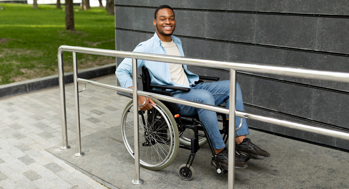 A happy man on a wheelchair enters the building on a ramp outdoors.